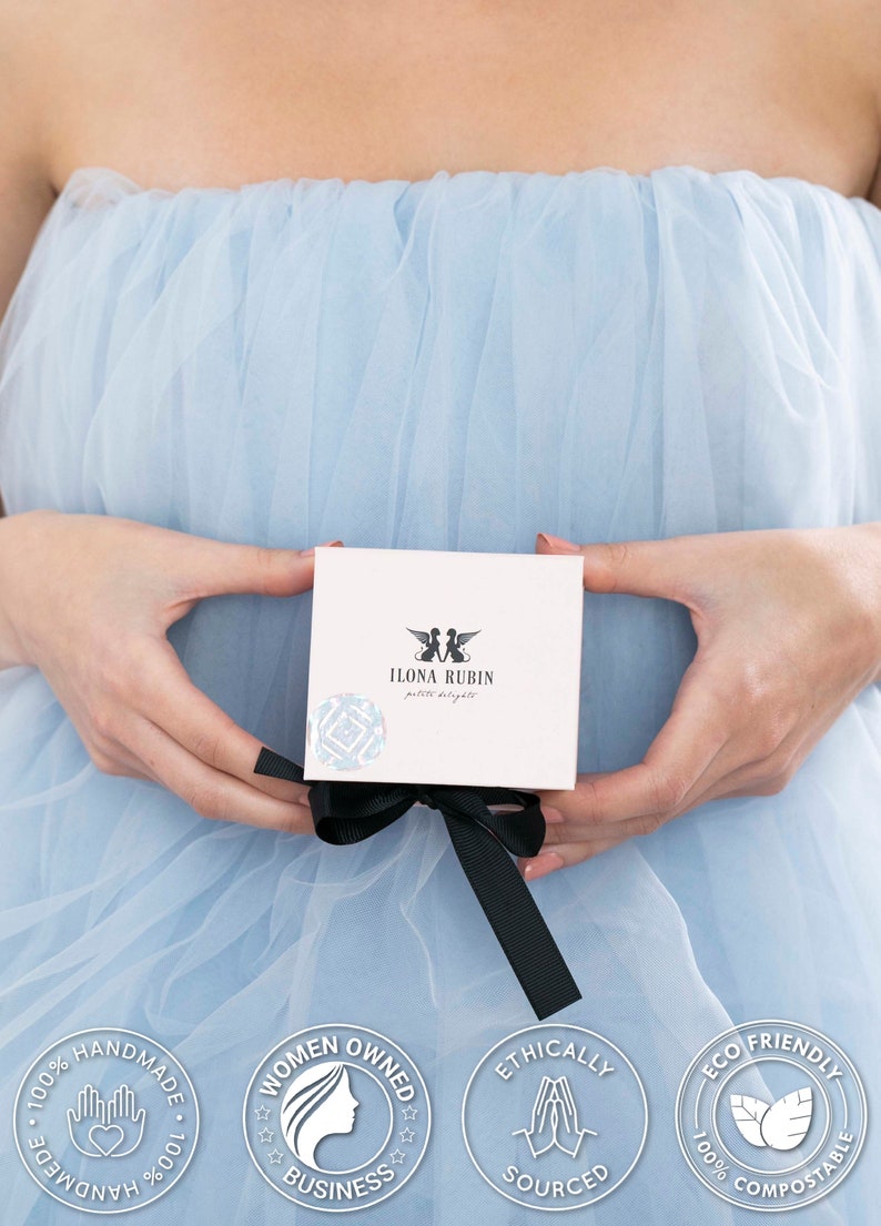 May include: A white box with a black ribbon tied around it. The box has a logo that says "ILONA RUBIN petite delights" and a pair of angels with wings. The box is being held by a person wearing a light blue tulle dress. There are four circular stamps on the dress that say "100% HANDMADE", "WOMEN OWNED BUSINESS", "ETHICALLY SOURCED", and "ECO FRIENDLY 100% COMPOSTABLE".