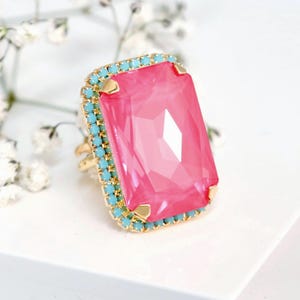 May include: A gold-toned ring featuring a large, rectangular, faceted hot pink center stone surrounded by a border of small, round, turquoise-colored crystals. The base of the ring is marked with the text ILONA RUBIN embellished with Swarovski crystals.