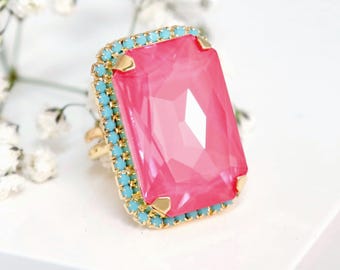 May include: A gold-toned ring featuring a large, rectangular, faceted hot pink center stone surrounded by a border of small, round, turquoise-colored crystals. The base of the ring is marked with the text ILONA RUBIN embellished with Swarovski crystals.