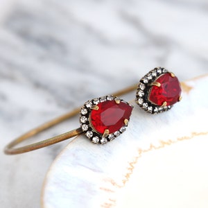 May include: A gold bangle bracelet with two teardrop-shaped red stones set in a clear crystal halo.