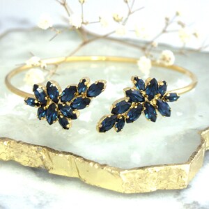 May include: A gold bangle bracelet with two blue crystal floral designs. The bracelet is on a gold and white marble surface.