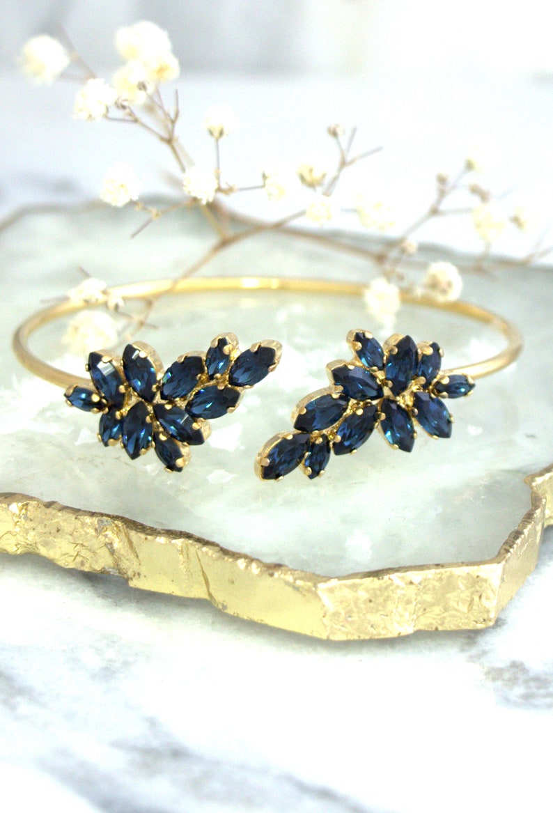 May include: A gold bangle bracelet with two blue crystal floral designs. The bracelet is on a gold and white marble surface.