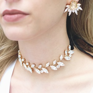 May include: A gold choker necklace with a delicate leaf design featuring clear crystal stones. The necklace is worn by a person with blonde hair and a white top.