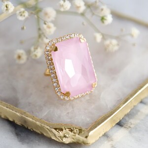 May include: A gold ring featuring a large, rectangular, faceted light pink stone set in the center. The stone is surrounded by a halo of small, clear, sparkling stones set into the gold band, which has four prongs holding the pink center stone.