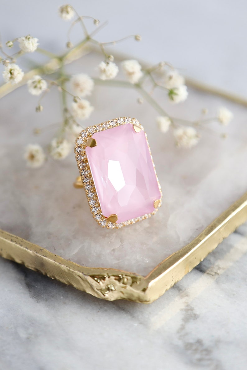 May include: A gold ring featuring a large, rectangular, faceted light pink stone set in the center. The stone is surrounded by a halo of small, clear, sparkling stones set into the gold band, which has four prongs holding the pink center stone.