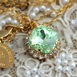 May include: A gold chain necklace with a light green crystal pendant. The pendant is surrounded by a halo of smaller clear crystals. The necklace is on a white lace background.