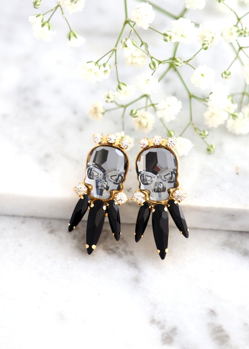 May include: A pair of gold tone earrings with black and clear crystal accents. The earrings feature a skull design with black crystal accents.