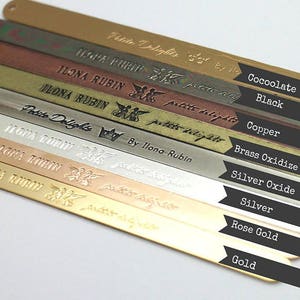 May include: A close-up of ten metal strips, each a different color, with the words "Petite Delights" and "Ilona Rubin" engraved on them. The colors are: gold, rose gold, silver, silver oxide, brass oxidize, copper, black, and cocoolate.