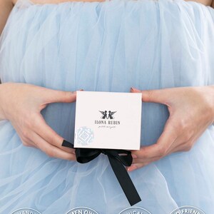 May include: A white box with a black ribbon is held against a light blue background. The box has the text "ILONA RUBIN petite delights" and a logo. The image also includes circular badges with text like "Handmade" and "Ethically Sourced".