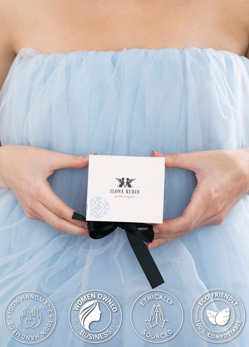 May include: A white box with a black ribbon is held against a light blue background. The box has the text "ILONA RUBIN petite delights" and a logo. The image also includes circular badges with text like "Handmade" and "Ethically Sourced".