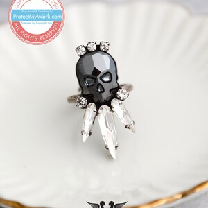 Puede incluir: Un anillo de calavera negro con detalles de cristal transparente. El anillo est&aacute; sobre un plato blanco con borde dorado.