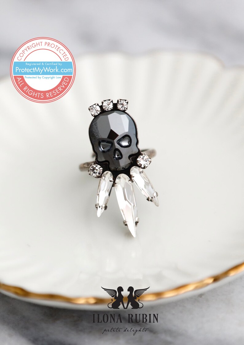 Puede incluir: Un anillo de calavera negro con detalles de cristal transparente. El anillo est&aacute; sobre un plato blanco con borde dorado.