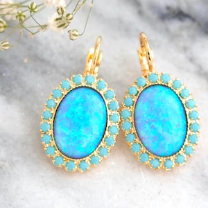 Op de afbeelding: Een paar goudkleurige oorbellen met hefboomsluiting, voorzien van een grote ovale blauwe steen met opaal-effect, omringd door een halo van kleine turquoise kristallen. Onderaan staat de tekst ILONA RUBIN petite delights embellished with Swarovski crystals.
