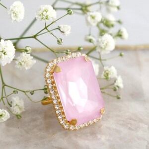 May include: A gold-toned ring featuring a large, rectangular, faceted light pink stone set in a frame of small, clear, sparkling crystals. The setting includes four gold-toned prongs at the corners. The brand name ILONA RUBIN is visible.