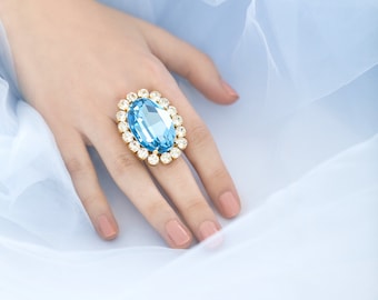 May include: A hand wearing a ring with a large blue oval-shaped gemstone surrounded by smaller clear gemstones.