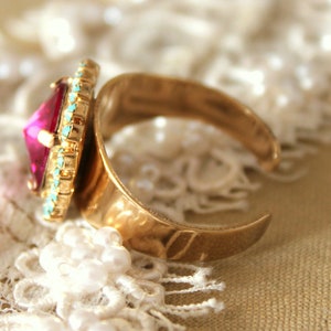May include: A gold ring with a large, faceted, pink gemstone surrounded by a turquoise and gold trim.