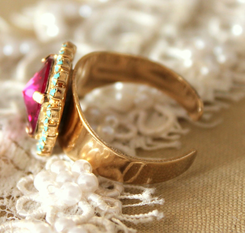 May include: A gold ring with a large, faceted, pink gemstone surrounded by a turquoise and gold trim.