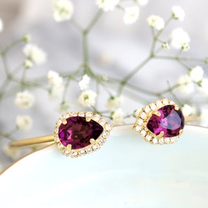 May include: A pair of gold earrings with pear-shaped purple gemstones surrounded by a halo of smaller clear gemstones.