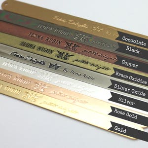 Op de afbeelding: Een close-up van tien metalen strips, elk met een andere afwerking, die de kleurvariaties van het metaal laten zien. De strips zijn gelabeld met het type metaal: Cocoolate, Zwart, Koper, Messingoxide, Zilveroxide, Zilver, Rosegoud en Goud.