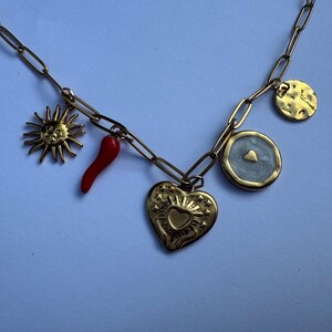 Pode incluir: Colar de pingentes dourado com um sol, pimenta vermelha, coração e pingentes circulares. O colar tem uma corrente de elos e é apresentado sobre um fundo azul claro. Os pingentes apresentam designs detalhados.