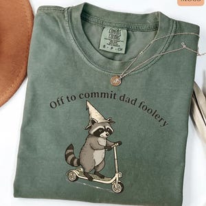 May include: Moss green t-shirt with the phrase "Off to commit dad foolery" and a cartoon raccoon riding a scooter. The raccoon wears a pointed hat. A silver necklace with a pendant is draped on the shirt.