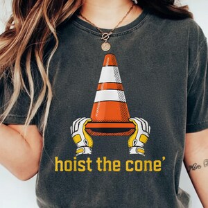 Pode incluir: T-shirt cinzenta escura com um gr&aacute;fico de um cone de tr&acirc;nsito segurado por m&atilde;os enluvadas, com o texto amarelo "hoist the cone". O cone &eacute; laranja e branco. A t-shirt tem gola redonda.