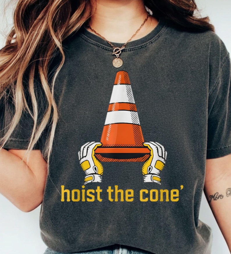 Pode incluir: T-shirt cinzenta escura com um gr&aacute;fico de um cone de tr&acirc;nsito segurado por m&atilde;os enluvadas, com o texto amarelo "hoist the cone". O cone &eacute; laranja e branco. A t-shirt tem gola redonda.