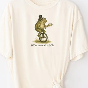 May include: Cream-colored t-shirt featuring a whimsical illustration of a frog riding a unicycle while holding a teacup. The frog wears a top hat. Text on the shirt reads "Off to cause a kerfuffle."