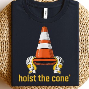 Pode incluir: Camiseta azul marinho com um gr&aacute;fico de um cone de tr&acirc;nsito laranja e branco segurado por m&atilde;os enluvadas. O texto "hoist the cone" est&aacute; impresso em amarelo abaixo. A camiseta &eacute; exibida em uma superf&iacute;cie tecida.