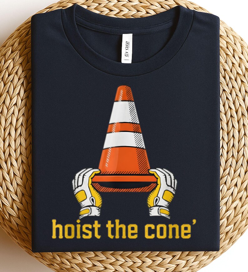 Pode incluir: Camiseta azul marinho com um gr&aacute;fico de um cone de tr&acirc;nsito laranja e branco segurado por m&atilde;os enluvadas. O texto "hoist the cone" est&aacute; impresso em amarelo abaixo. A camiseta &eacute; exibida em uma superf&iacute;cie tecida.
