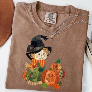 Puede incluir: Una camiseta marrón de manga corta con un diseño bordado tipo parche de un espantapájaros con sombrero negro y un cuervo, rodeado de calabazas estampadas naranjas y verdes. La etiqueta interior dice COMFORT COLORS S P CH.