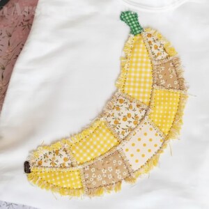 May include: White sweatshirt with a patchwork banana design. The banana is made from various yellow and tan fabrics, including gingham, floral, and polka dot patterns. It has a green stem and a frayed edge.
