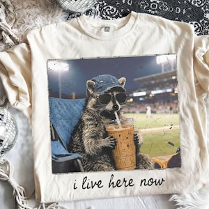Può includere: T-shirt color crema con un procione con occhiali da sole e berretto da baseball, che beve da un bicchiere. L'immagine ha uno sfondo di stadio di baseball e il testo "i live here now".