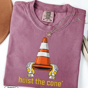 Pode incluir: T-shirt cor de baga com um gr&aacute;fico de um cone de tr&acirc;nsito segurado por m&atilde;os enluvadas e o texto "hoist the cone". A camisa tem uma etiqueta Comfort Colors e um pequeno colar com pingente redondo. A camisa est&aacute; dobrada.