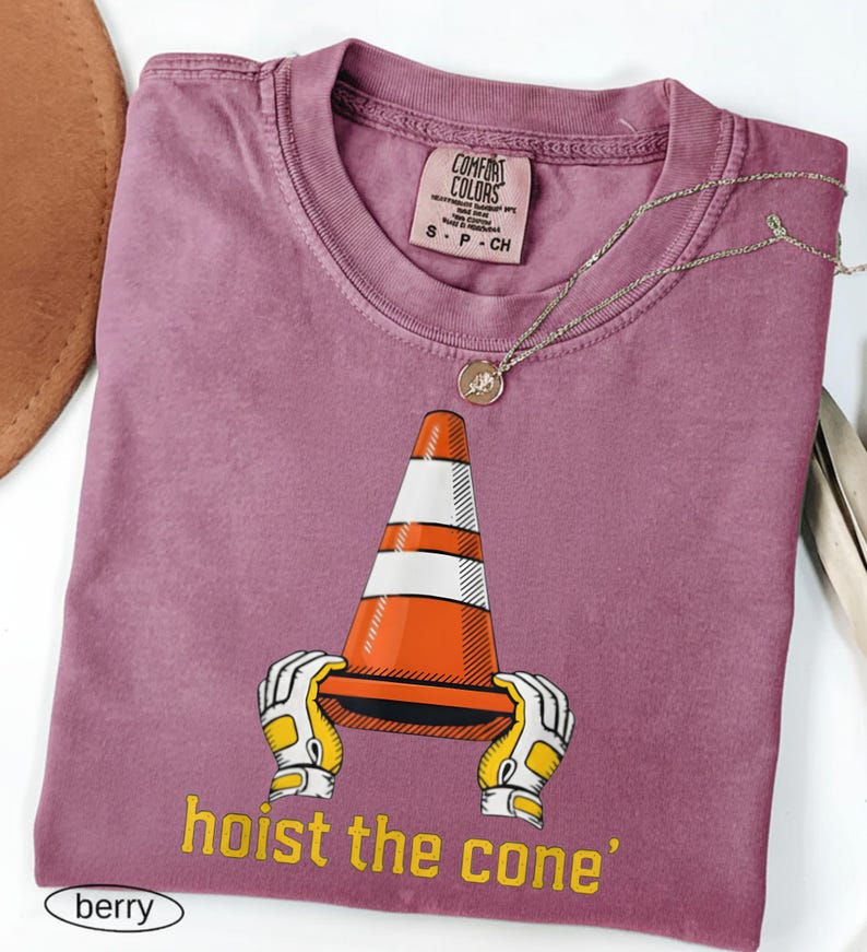 Pode incluir: T-shirt cor de baga com um gr&aacute;fico de um cone de tr&acirc;nsito segurado por m&atilde;os enluvadas e o texto "hoist the cone". A camisa tem uma etiqueta Comfort Colors e um pequeno colar com pingente redondo. A camisa est&aacute; dobrada.