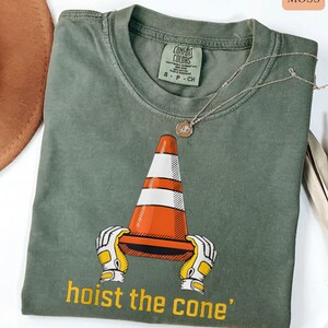 Pode incluir: T-shirt verde musgo com um gr&aacute;fico de um cone de tr&acirc;nsito laranja e branco segurado por m&atilde;os enluvadas. O texto "hoist the cone" est&aacute; impresso em amarelo. Um colar de prata com um pingente est&aacute; sobre a camisa.