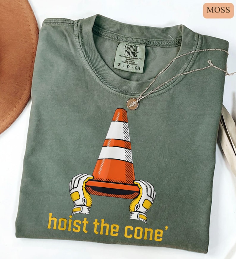 Pode incluir: T-shirt verde musgo com um gr&aacute;fico de um cone de tr&acirc;nsito laranja e branco segurado por m&atilde;os enluvadas. O texto "hoist the cone" est&aacute; impresso em amarelo. Um colar de prata com um pingente est&aacute; sobre a camisa.