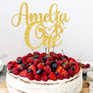 May include: A celebration cake with white frosting and a topping of fresh berries, such as strawberries, raspberries, and blueberries. A gold glitter cake topper says "Amelia is One". The cake is on a wooden board.