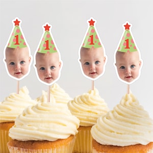 May include: Cupcakes with white frosting and baby face toppers wearing party hats with the number 1. The hats are green with pink polka dots and a red star on top. The cupcakes are for a first birthday celebration.