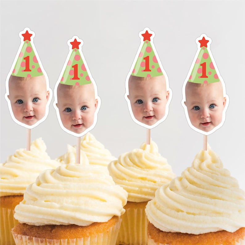 May include: Cupcakes with white frosting and baby face toppers wearing party hats with the number 1. The hats are green with pink polka dots and a red star on top. The cupcakes are for a first birthday celebration.