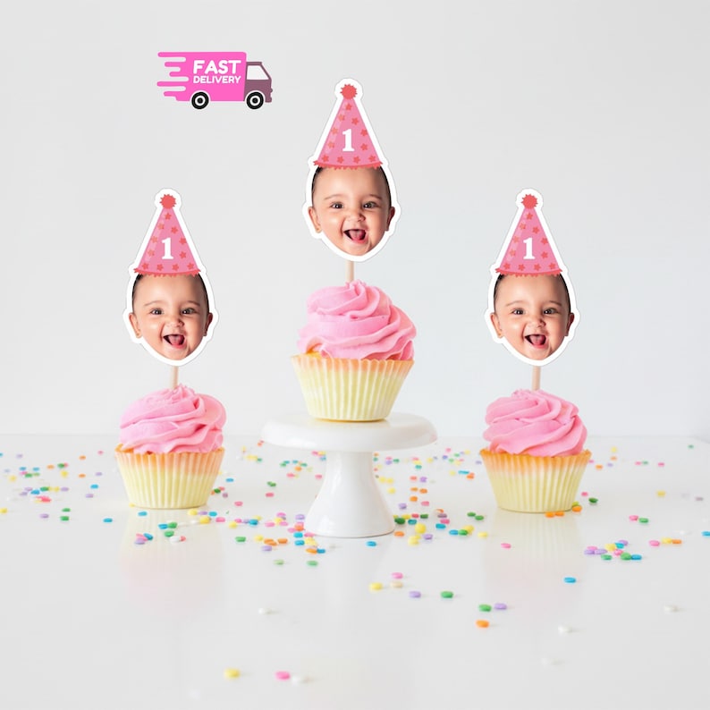 May include: Pink frosted cupcakes with baby face toppers wearing party hats with the number 1. A white cake stand holds one cupcake. A pink delivery truck graphic is in the background. Colorful sprinkles are scattered around the cupcakes.