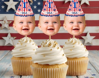 Könnte beinhalten: Drei Cupcakes mit weißem Zuckerguss und goldenen Streuseln, jeweils gekrönt von einem Babygesicht und einer patriotischen Partyhütchen. Die Hüte sind rot, weiß und blau mit dem Wort "USA" und der Zahl "1". Der Hintergrund zeigt eine amerikanische Flagge.