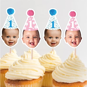 May include: Cupcakes topped with decorative picks featuring smiling baby faces and party hats. The hats are decorated with a number 1, confetti, and a pom-pom. The cupcakes have a swirl of yellow frosting.