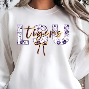 May include: White sweatshirt with the text "LSU Tigers" in purple and gold. The letters "LSU" are filled with a floral pattern in purple and white. A gold bow and ribbon accent the design.