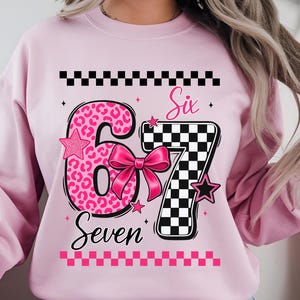 Könnte beinhalten: Rosa Sweatshirt mit einem grafischen Design mit den Zahlen "67", den Wörtern "Six" und "Seven". Die Zahl "6" hat einen rosa Leopardenmuster, und die Zahl "7" hat ein schwarz-weißes Schachbrettmuster. Eine rosa Schleife und Sterne sind ebenfalls enthalten.