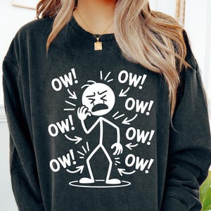May include: Dark gray sweatshirt with a white graphic of a stick figure in pain, clutching their face. The word "OW!" is printed around the figure in multiple directions, emphasizing the distress. A casual, humorous design.