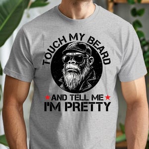 Puede incluir: Camiseta gris claro con una ilustración en blanco y negro de un simio barbudo con gafas de sol y gorra. El texto dice "TOUCH MY BEARD AND TELL ME I'M PRETTY" en un diseño circular.