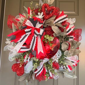 Può includere: Una ghirlanda decorativa con accenti rossi, bianchi e neri. La ghirlanda include un grande cuore rosso, un fiocco con pois neri e vari nastri e verde. La ghirlanda è progettata per la decorazione della porta.