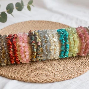 Pode incluir: Uma coleção de pulseiras de lascas de pedras preciosas coloridas, exibidas em um tapete tecido. As pulseiras apresentam uma variedade de cores, incluindo roxo, marrom, vermelho, rosa, azul e verde. Circunferência de aproximadamente 18 cm.