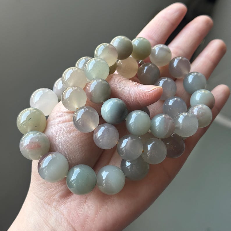 May include: A bracelet of large, spherical beads in a palette of green, grey, and white. The beads are polished and have a shiny surface. The bracelet is held in a hand, highlighting the natural colour and texture variations of the stones.